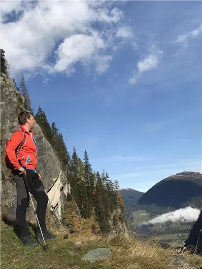 A person is standing on a hiking trail and is gazing at the view of the mountains. The sky is blue and covered with some clouds.