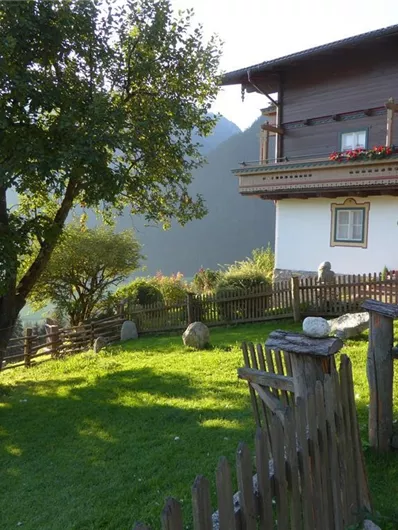 Ein gemütliches Holzhaus mit einem Balkon und bunten Blumen. Der Garten ist grün und von einem Holzzaun umgeben, mit einem Blick auf die Berge.