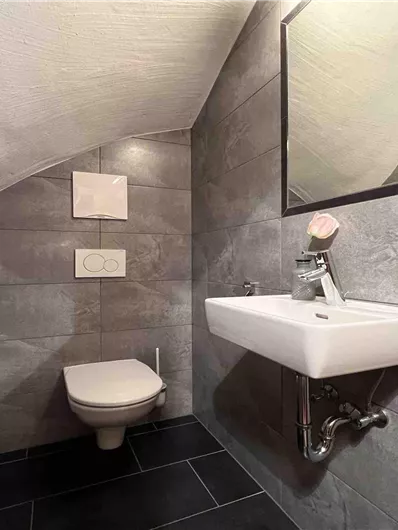 Ein modernes Badezimmer mit grauen Fliesen und einem Waschbecken. Die Wand ist schräg und es gibt einen Spiegel über dem Waschbecken.