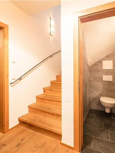 Ein heller Flur mit Holztreppe und zwei offenen Türen. Links führt die Treppe nach oben, rechts befindet sich ein kleines Badezimmer.