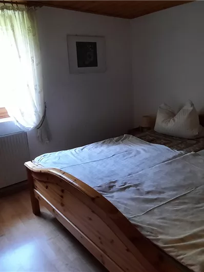 Een gezellige slaapkamer met een groot bed en lichte gordijnen. De wanden zijn eenvoudig en de ruimte heeft veel daglicht.