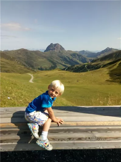 Een klein jongetje zit op een houten plank en kijkt glimlachend in de camera. Op de achtergrond zijn groene heuvels en bergen onder een blauwe lucht te zien.