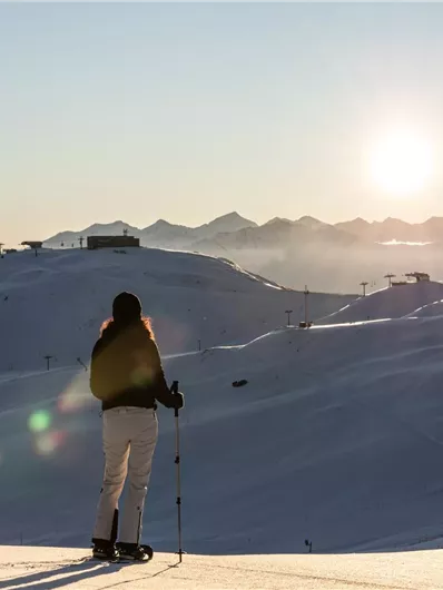 Eine Person steht auf einem verschneiten Hügel und betrachtet die aufgehende Sonne. Im Hintergrund sind schneebedeckte Berge und Skilifte zu sehen.