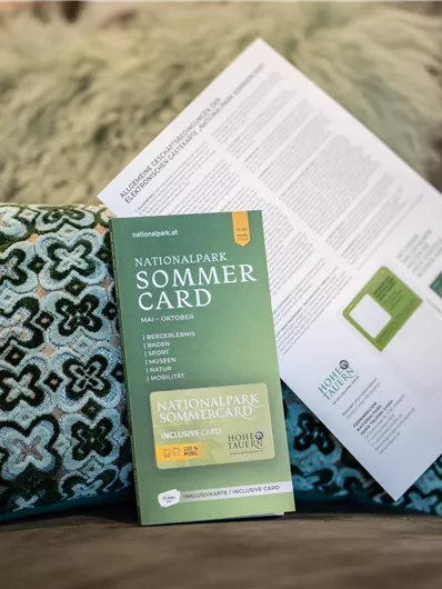 Een zomermanifest ligt op een sofa naast een decoratief kussen. Op de achtergrond is een informatieblad te zien.