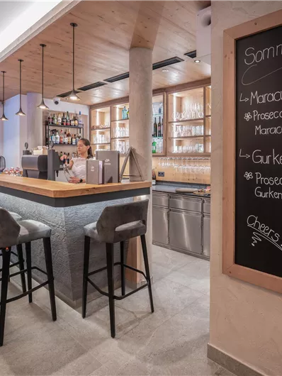 Een moderne bar met houtdecor en hoge stoelen. Aan de muur hangt een bord met drankspecials.