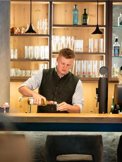Een bartender bereidt een drankje voor aan een stijlvolle bar. Op de achtergrond zijn talloze flessen en glazen te zien.
