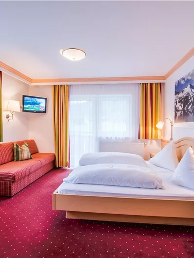 Ein gemütliches Hotelzimmer mit zwei Betten und einem Sofa. Große Fenster sorgen für viel Tageslicht und eine angenehme Atmosphäre.