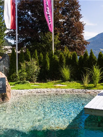 Ein stilvoller Außenbereich mit einem glitzernden Pool und bequemen Gartenmöbeln. Im Hintergrund sind grüne Bäume und majestätische Berge zu sehen.