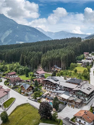 Eine malerische Berglandschaft mit einem kleinen Dorf und umliegenden Wäldern. Die Häuser sind verstreut und der Himmel ist teilweise bewölkt.