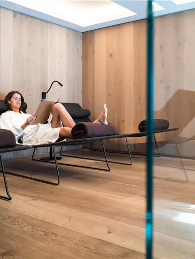 Een ontspannend wellnessgebied met houten bekleding. Een vrouw zit in een badjas op een ligstoel en leest.