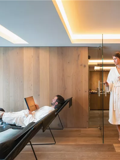 A modern spa with wooden paneling. One person is relaxing on a lounger, while another is standing in a bathrobe.