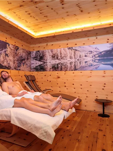 A cozy wellness room with wooden paneling. Two people are relaxing on loungers and enjoying the calm atmosphere.