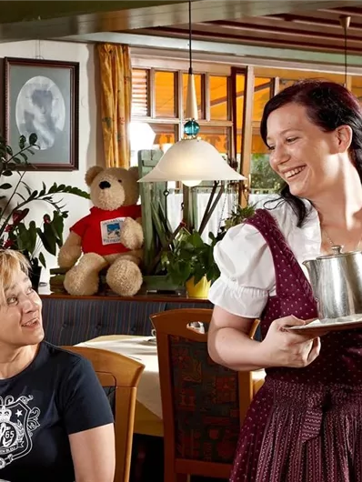 Een serveerster brengt een kan naar een vrouw aan tafel. Op de achtergrond is een teddy en een uitnodigende restaurantatmosfeer te zien.