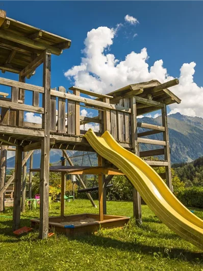 Een houten speelplaats met een glijbaan staat in de tuin. Op de achtergrond zijn bergen en een blauwe lucht met wolken te zien.
