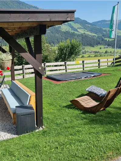 Ein einladender Garten mit einer Holzveranda und Liegestühlen. Im Hintergrund sind grüne Hügel und ein Trampolin zu sehen.
