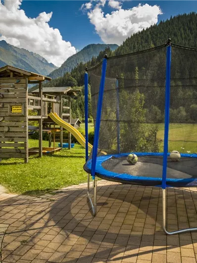 Een speelplaats met een trampoline en een houten speelstructuur. De omgeving is groen met bergen en een heldere lucht op de achtergrond.