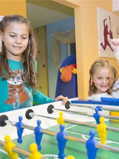 Twee kinderen spelen enthousiast tafelvoetbal. Het meisje rechts is blij en steekt haar hand op, terwijl het andere meisje geconcentreerd speelt.