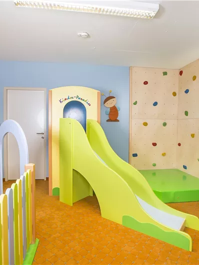 Ein bunten Spielzimmer mit einer Rutsche und einer Kletterwand. Die Wände sind in hellen Farben gehalten und es gibt eine kleine Spielzeugumrandung.