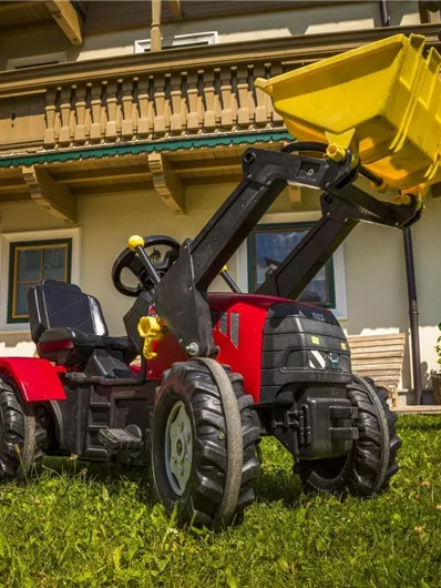 Een rood speelgoedvoertuig met een gele schep staat op het gras. Op de achtergrond is er een gebouw met een balkon te zien.
