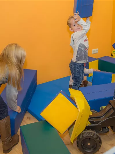 Drei Kinder spielen in einem bunten Raum mit Schaumstoffblocks. Eines der Kinder baut mit den Blöcken, während die anderen auf einem Fahrzeug fahren.