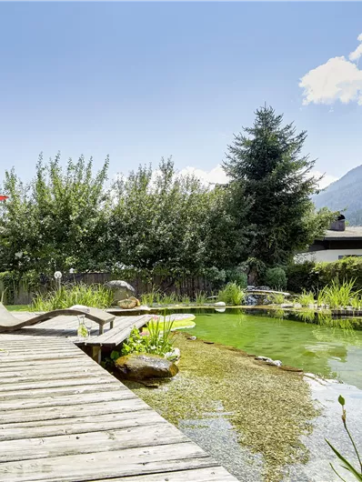 Een idyllische tuin met een houten pad dat naar een heldere vijver leidt. Op de achtergrond zijn bomen en bergen te zien, terwijl een rode parasol schaduw biedt.