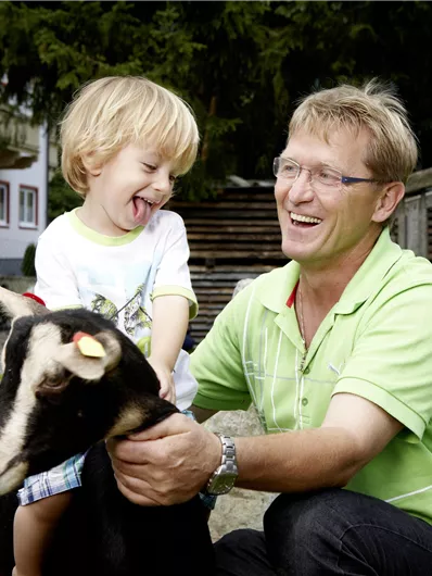 Ein fröhliches Kind und ein Mann lachen zusammen, während sie eine Ziege betreuen. Im Hintergrund sind ein Holzsteg und Bäume sichtbar.
