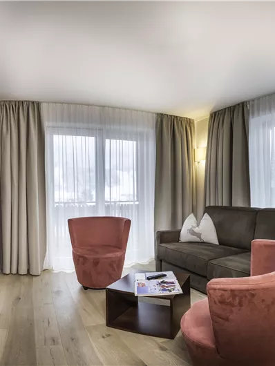 A cozy living room with a sofa, two red armchairs, and a table. Large windows provide plenty of light and a modern look.