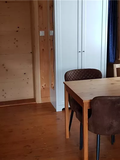 Ein gemütlicher Wohnraum mit einer kleinen Küchenzeile und einem Esstisch. Die Wände sind aus Holz und es gibt Fenster, die natürliches Licht hereinlassen.