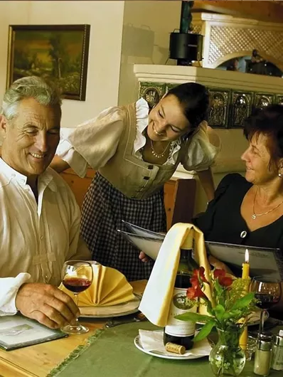 Een gezellig restaurant met een oudere man die glimlacht terwijl hij met een serveerster praat. Een vrouw zit aan tafel en kijkt ook glimlachend naar de menu's.