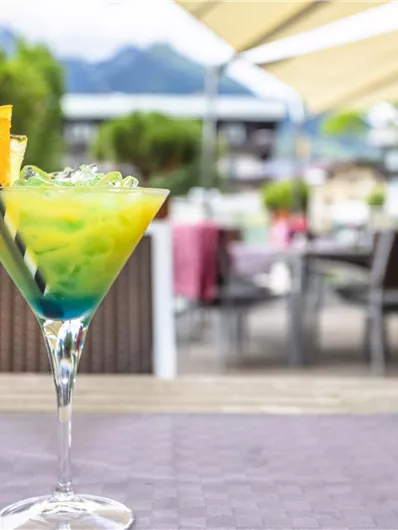 A colorful cocktail with pineapple and vibrant layers is sitting on a table. In the background, several seating areas and a green environment can be seen.