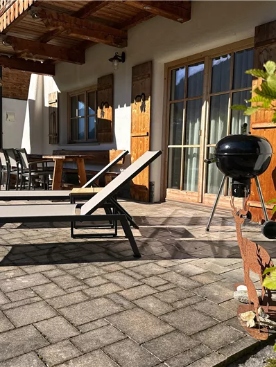 Eine sonnige Terrasse mit Liegen und einem Grill. Im Hintergrund stehen ein Tisch und Stühle, umgeben von einem gepflegten Garten.