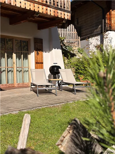 Eine gemütliche Terrasse mit zwei Liegen und einem Grill. Im Hintergrund sind Holzwände und grünes Gras zu sehen.