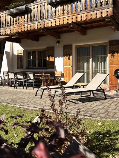 Eine gemütliche Terrasse mit Liegen und einem Grill. Im Hintergrund steht ein Holzbalcony und es gibt einen gepflegten Garten.