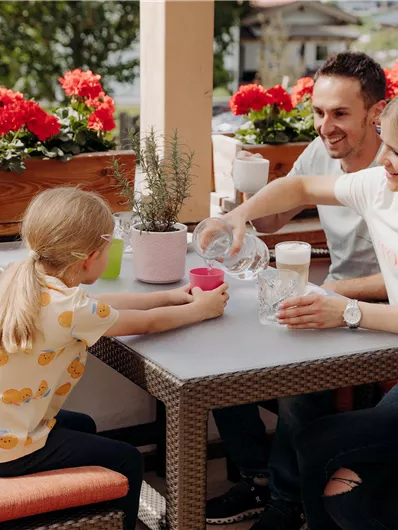 Eine Familie sitzt an einem Tisch im Freien und genießt Getränke. Im Hintergrund blühen rote Blumen in Kästen.
