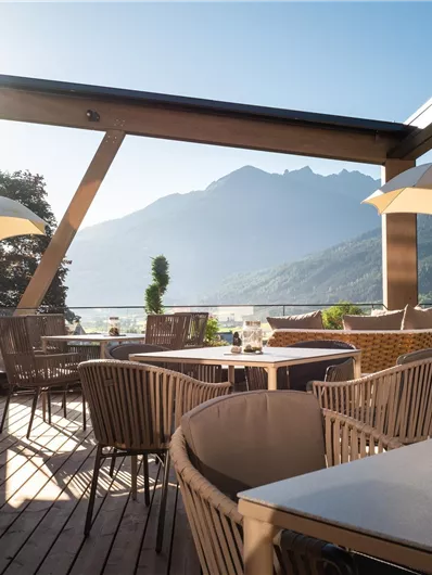 Eine einladende Terrasse mit gemütlichen Stühlen und Sonnenschirmen. Im Hintergrund erheben sich malerische Berge unter einem klaren Himmel.