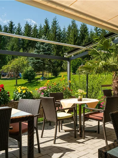 Eine angenehme Terrasse mit Tischen und Stühlen, umgeben von bunten Blumen. Im Hintergrund sind Bäume und ein klarer Himmel zu sehen.