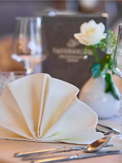 Een elegante eettafel met gevouwen servetten en glazen. Op de achtergrond is een bloemenarrangement en een menukaart te zien.