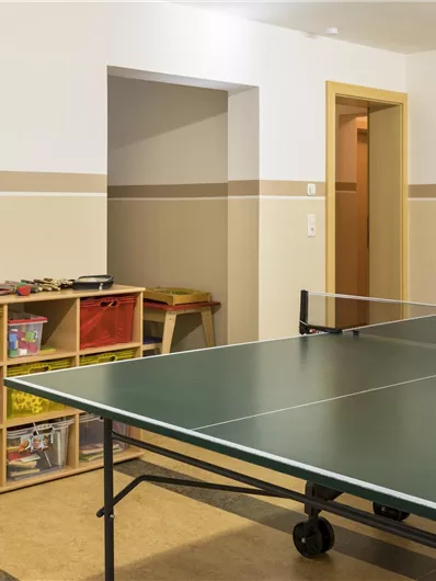 Een speelruimte met een tafeltennistafel en kleurrijke speelgoedopslag. De muren zijn licht geschilderd en er is toegang tot een andere kamer.