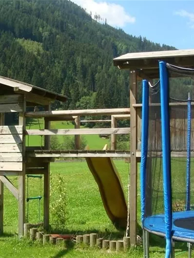 Een speeltuin met een houten speelstructuur, een glijbaan en schommels. Op de achtergrond zijn groene weilanden en bergen te zien.