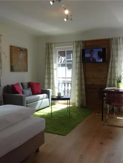 Ein modernes Zimmer mit einem gemütlichen Bett und einer Sitzecke. Es gibt große Fenster, die viel Licht hereinlassen, sowie einen Fernseher und einen kleinen Tisch.