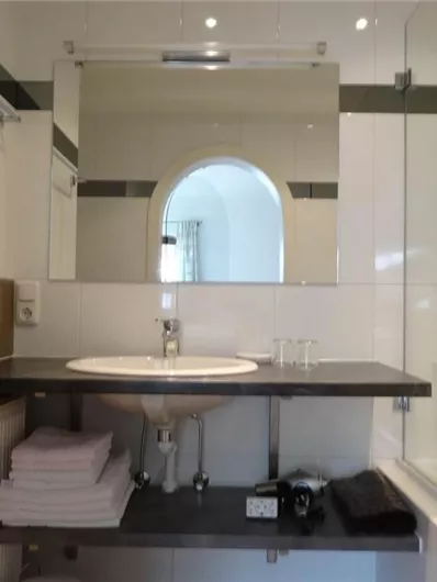 Ein modernes Badezimmer mit einem Waschbecken, Spiegel und Fön. Auf einem Regal liegen frische Handtücher und eine Badewanne ist sichtbar.