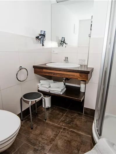 Ein modernes Badezimmer mit einer Dusche, Waschbecken und Toilette. Die Einrichtung ist einfach und funktional, mit hellen Fliesen und einem Holzregal.