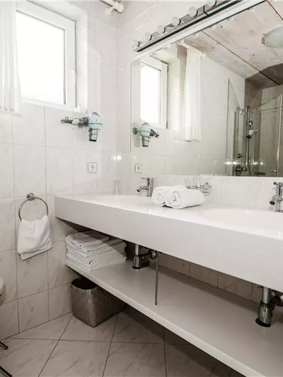 Een modern badkamers met lichte tegels en een grote spiegel. Er is een wastafel, handdoeken en een kruk.