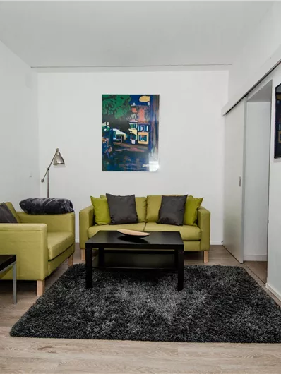 Ein modernes Wohnzimmer mit hellen, grünen Sofas und einem schwarzen Tisch. An der Wand hängt ein Bild, und ein Fernseher ist an der Seite montiert.