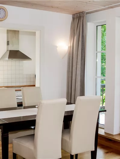 Eine moderne Küche mit einem Esstisch und weißen Stühlen. Große Fenster bieten Blick auf die Terrasse und den Garten.