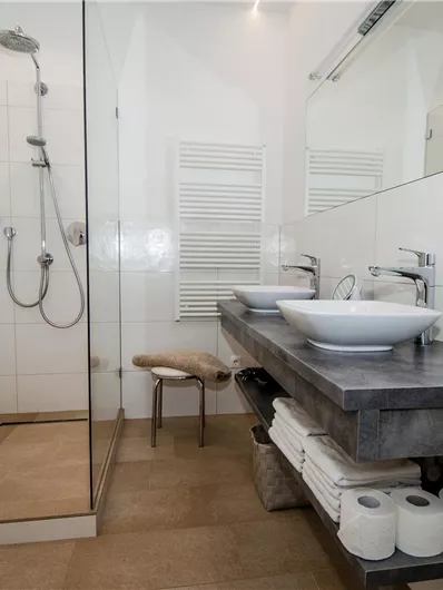 Ein modernes Badezimmer mit Dusche und zwei Waschbecken. Die Wände sind hell gefliest und es gibt einen Holzunterbau für Handtücher und Toilettenpapier.