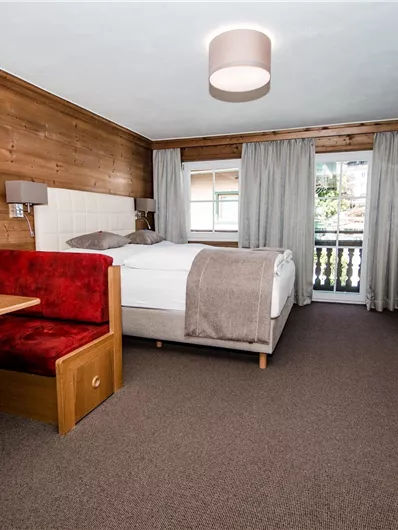 Een gezellige hotelkamer met houten panelen, een tweepersoonsbed en een balkon. Er is een klein tafeltje met rode stoelen en een televisie aan de muur.