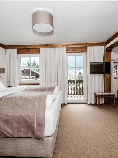 Een gezellige hotelkamer met een groot bed en uitzicht op de bergen. De kamer is licht en uitnodigend, met een houten tafel en stoelen, evenals een televisie.