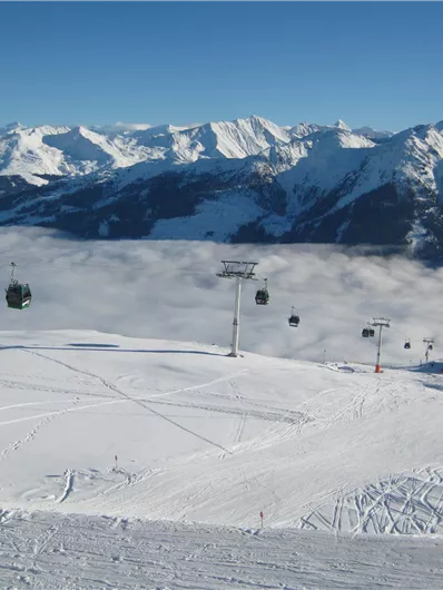 Eine schneebedeckte Berglandschaft mit Skiliften und majestätischen Bergen im Hintergrund. Die Wolken liegen tief im Tal und schaffen eine beeindruckende Aussicht.