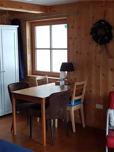 Ein gemütlicher Wohnraum mit Holzwänden, einem Esstisch und Stühlen. Im Hintergrund sind ein Schrank und ein Fernseher zu sehen.